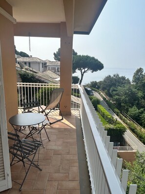 Ein Balkon mit Tisch und Stühlen, einem weißen Geländer und Blick auf eine Straße sowie Grünflächen.