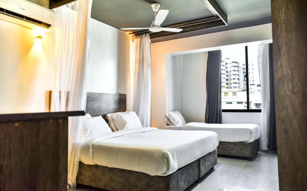 Une chambre d’hôtel avec deux lits, un ventilateur de plafond et une vue sur la ville grâce à de grandes fenêtres.