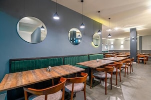 Un restaurante con mesas de madera y asientos tapizados verdes, luces colgantes y espejos redondos en las paredes.