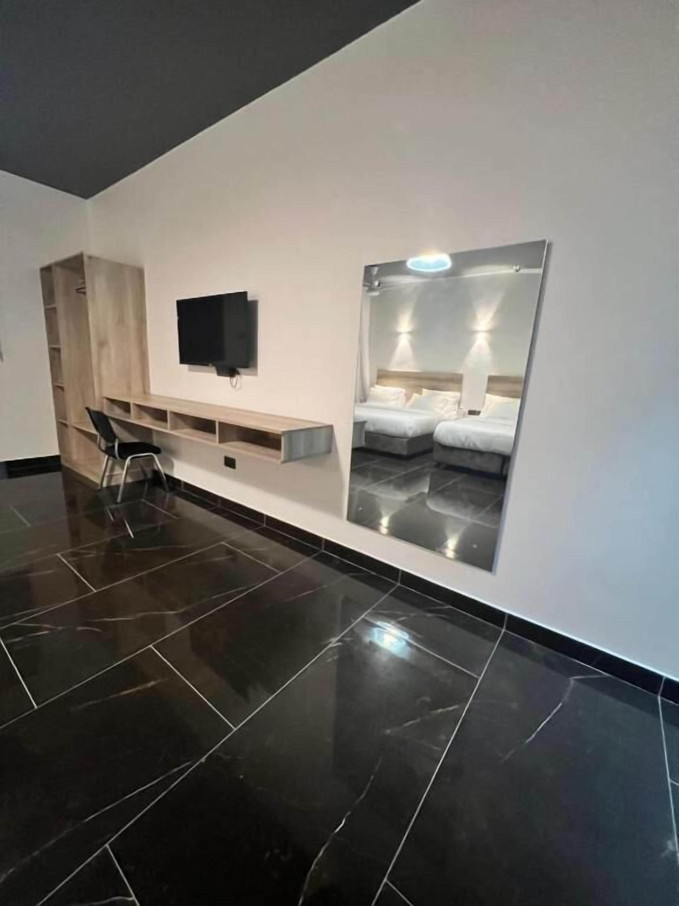 A modern hotel room with a large mirror reflecting a bed, a flat-screen TV mounted on the wall, and a wooden wardrobe with a built-in shelf.