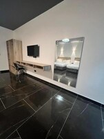 Una habitación de hotel moderna con un espejo grande que refleja una cama, un televisor de pantalla plana montado en la pared y un armario de madera con estante incorporado.