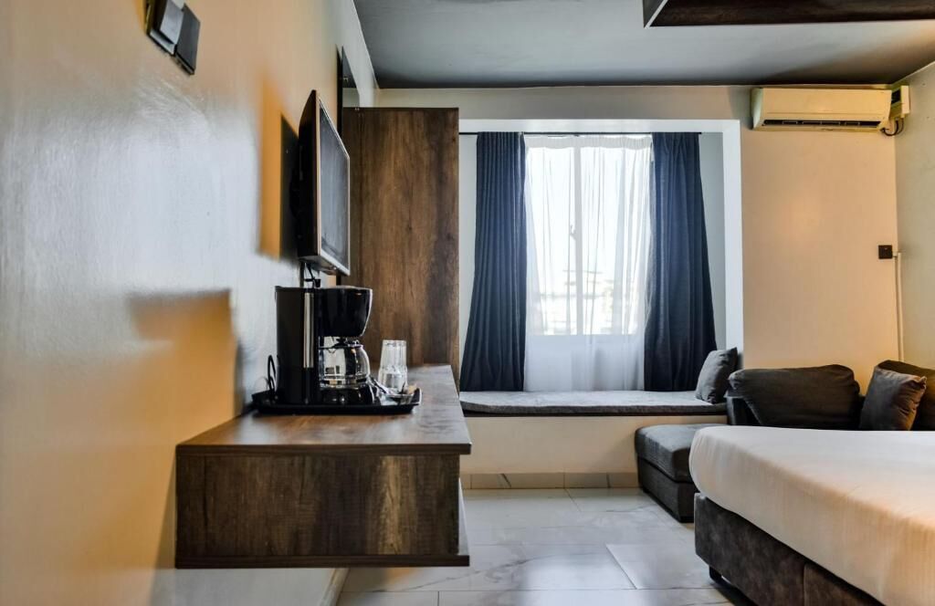 A hotel room with a bed, a small seating area by the window, a coffee station, and a flat-screen TV mounted on the wall.