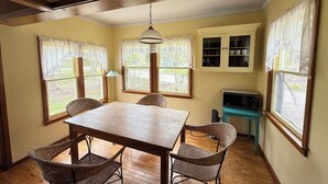 Un comedor con mesa de madera y sillas de mimbre, lámpara colgante y ventanas con cortinas.
