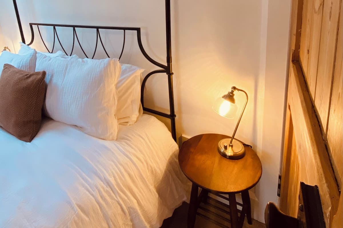 Una cama con dosel con ropa de cama blanca y una almohada marrón, una pequeña mesa de madera con una lámpara y una puerta de madera.