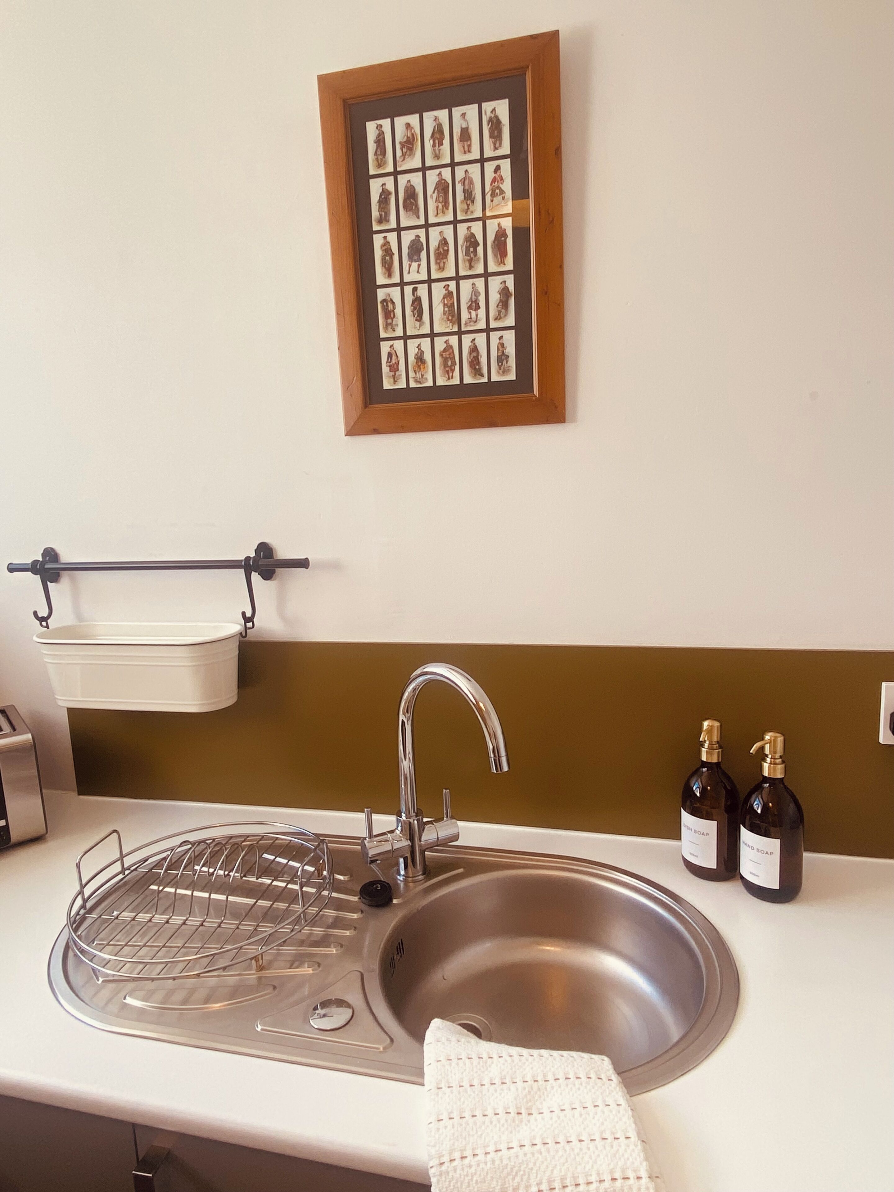 A kitchen with a stainless steel sink, a hanging soap dispenser, a framed picture on the wall, and a white towel on the counter.