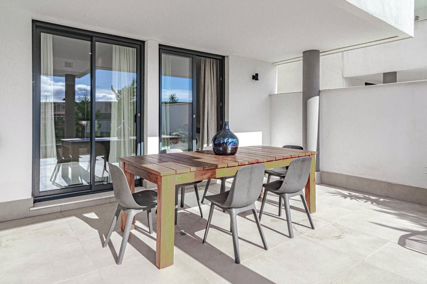 A wooden table with four chairs, a vase, and a glass partition in a sunlit outdoor area.