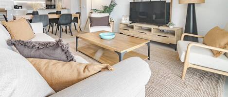 A modern living room with a sofa, coffee table, and television. There is a dining area with chairs and a table.