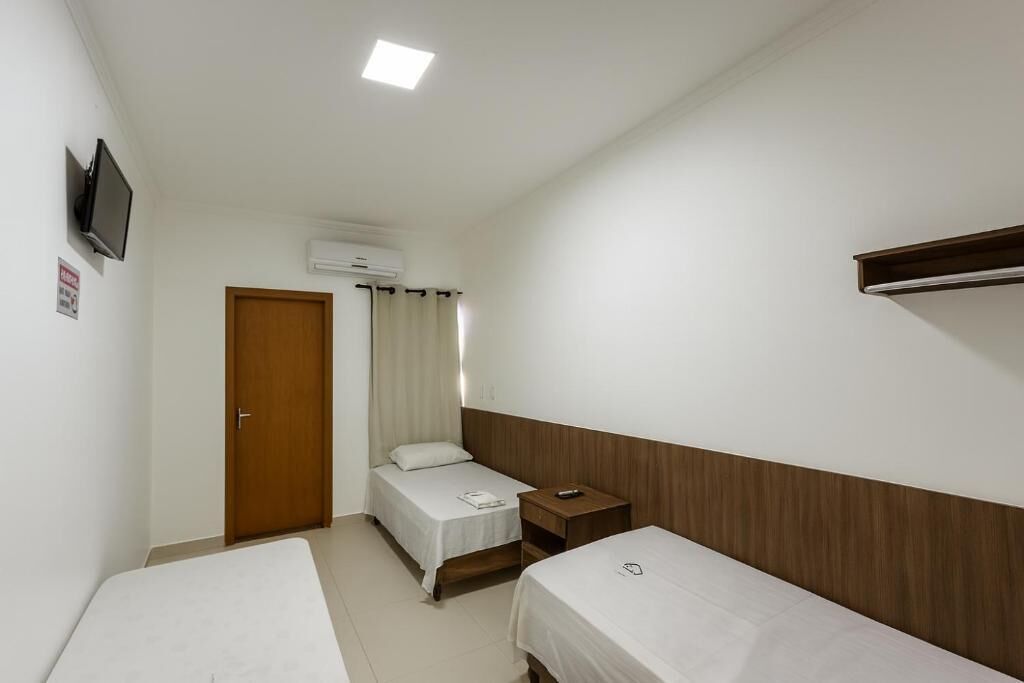 A small, clean room with two single beds, a wooden nightstand, a TV mounted on the wall, and a door with a curtain.