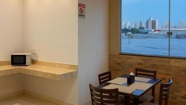 A dining area with a table and chairs, a microwave on a counter, and a view of a cityscape through a large window. - Nova Jerusalém Hotel (Goiânia)