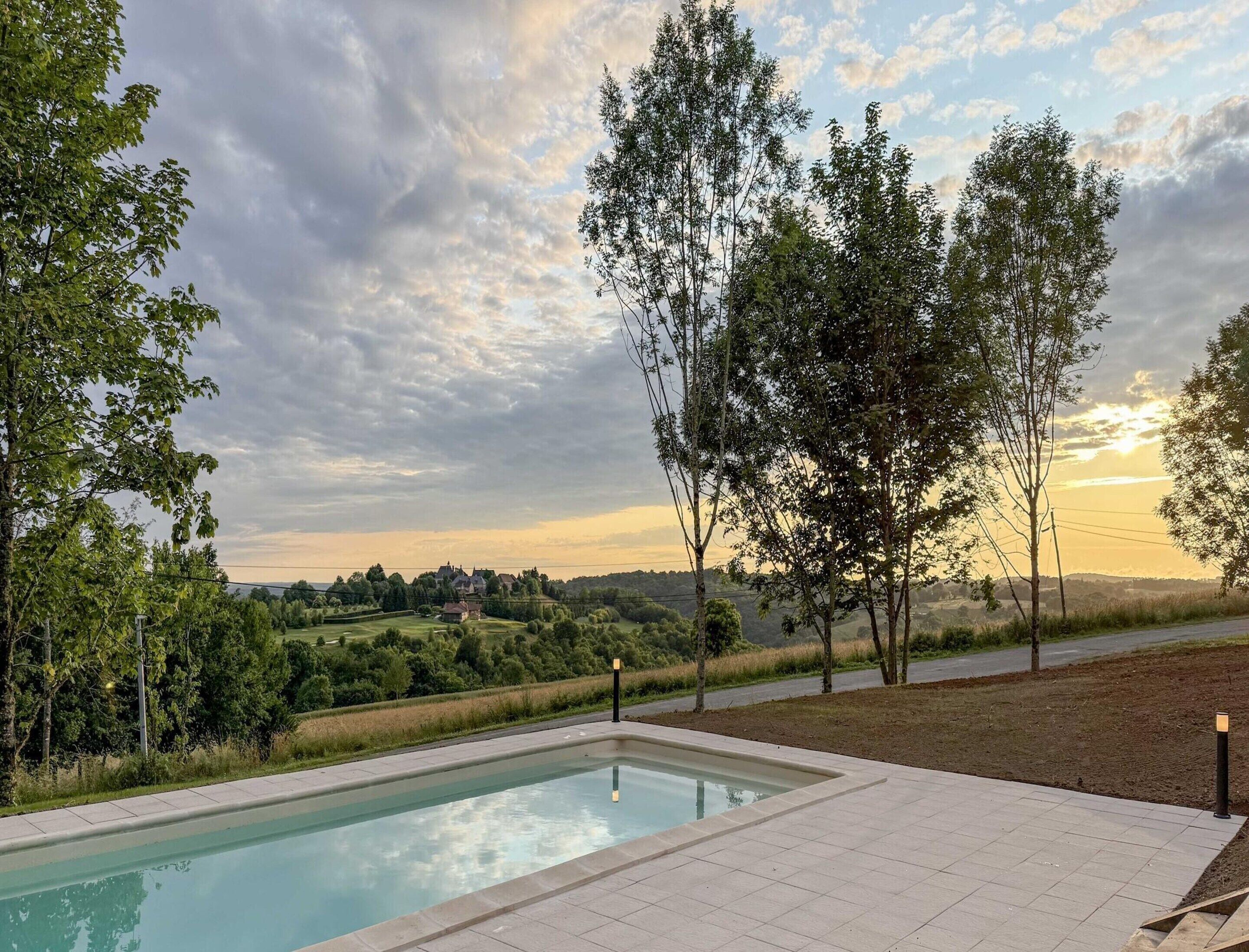 Une piscine avec une terrasse carrelée, entourée d’arbres et offrant une vue sur un paysage rural avec des maisons visibles au loin.