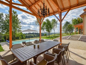 A covered outdoor area with a pool, dining table, and chairs, overlooking a scenic landscape. - Elegant 10-guest Villa With Pool & Castle Views (Saint-Medard-d'Excideuil)