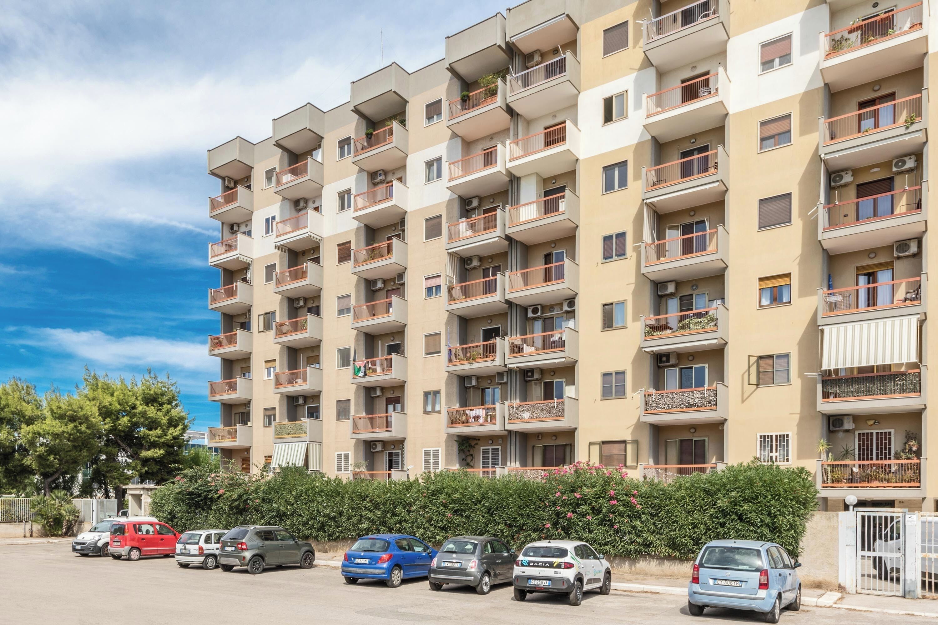 A multi-story residential building with balconies, a parking area with several cars, and a clear sky.