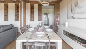 Un comedor moderno con una mesa blanca dispuesta para seis personas, rodeada de sillas. Un sofá gris está situado a la izquierda, y un armario blanco con una obra de arte de rosas a la derecha.