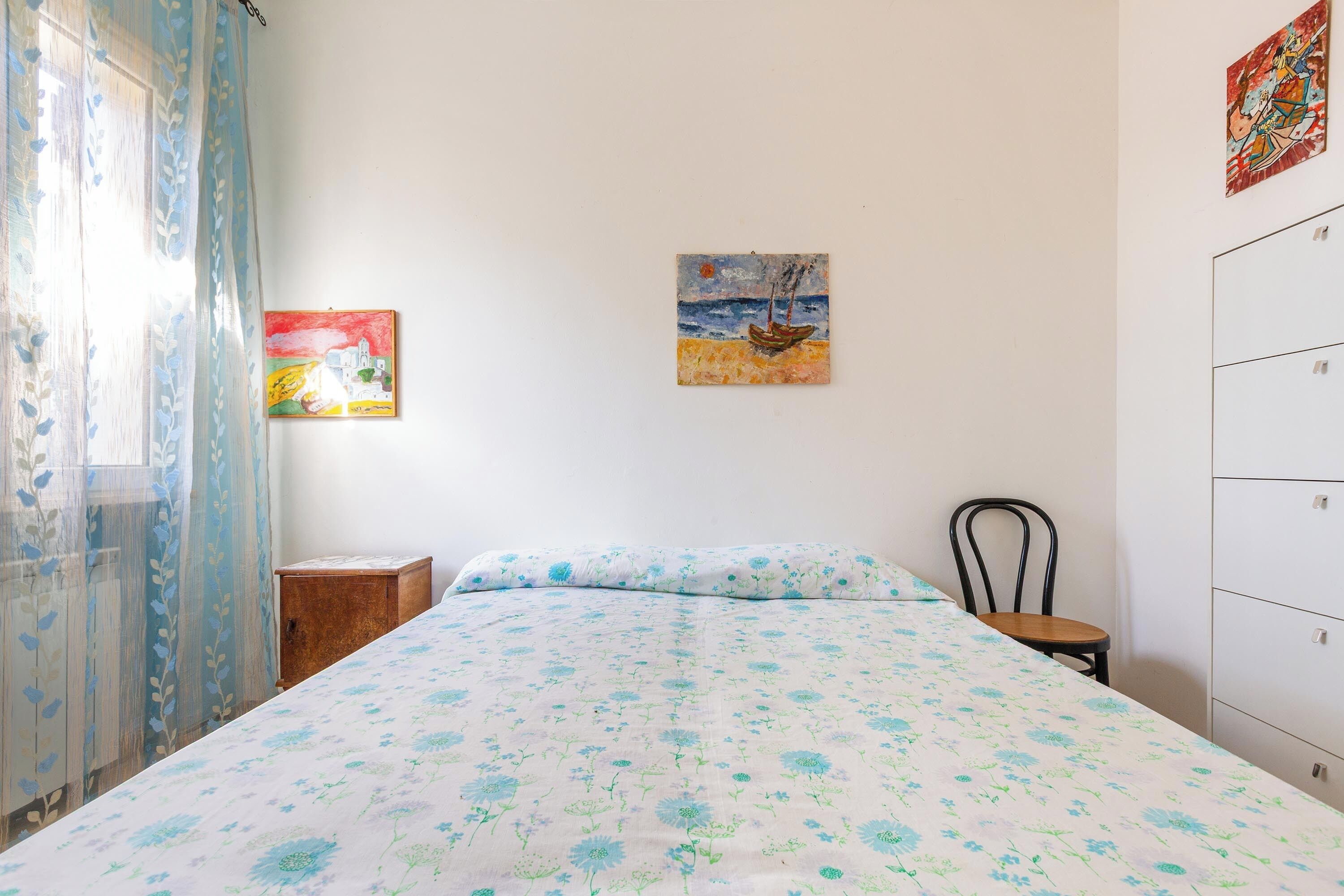 A bedroom with a bed, a chair, a small wooden cabinet, and two paintings on the wall.