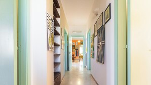 Um corredor com escada à esquerda, paredes decoradas com quadros, e uma porta no final que dá para um quarto com cadeira e mesa.