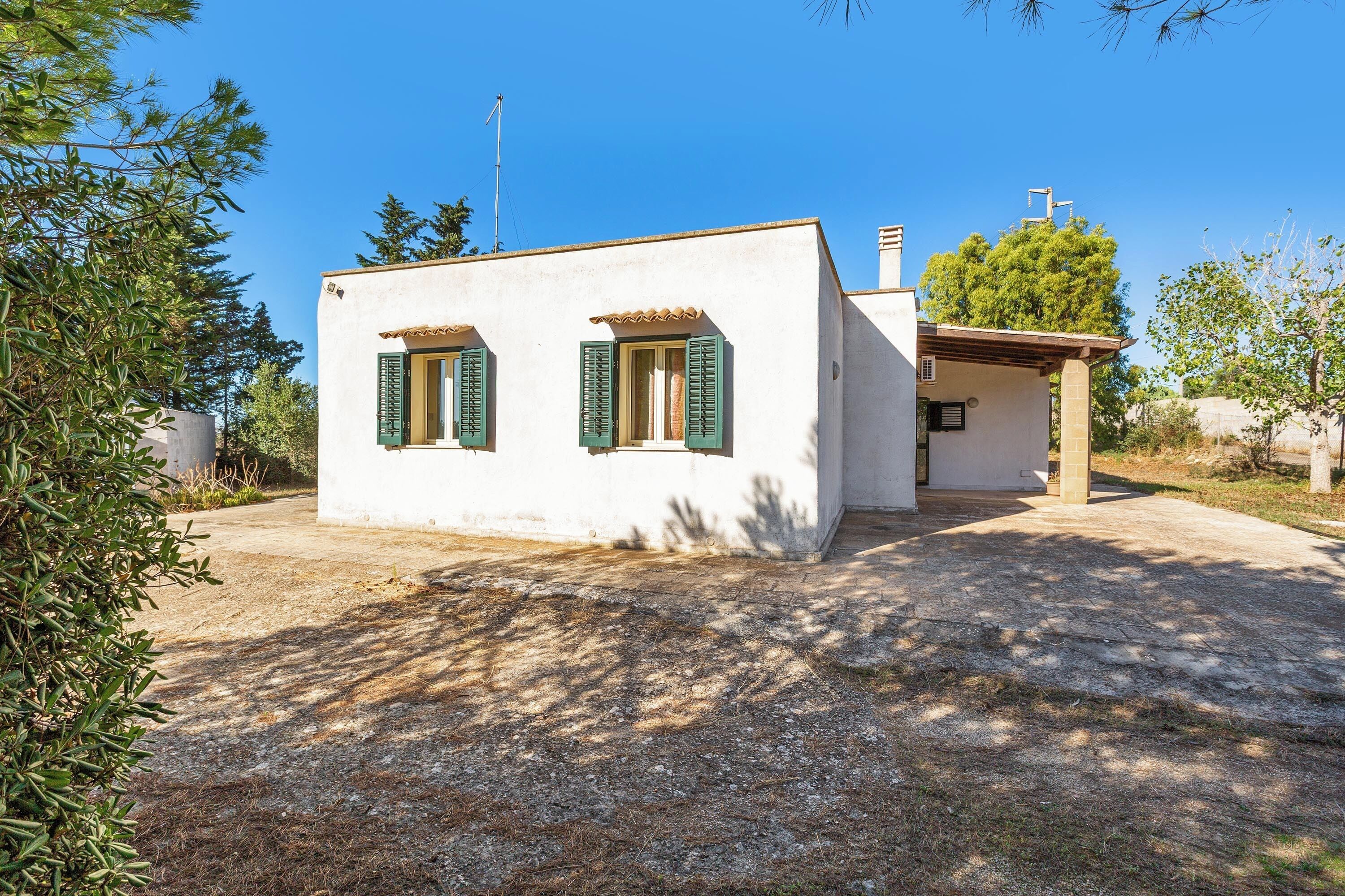 A small, single-story house with a white exterior and green shutters, surrounded by trees and a clear sky.