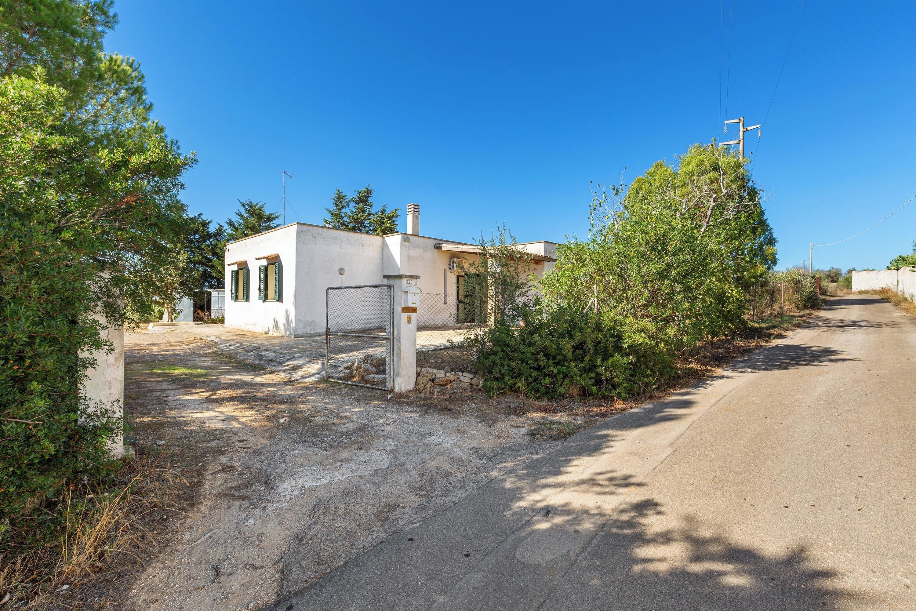 A small, single-story building with a concrete exterior, a metal gate, and a paved driveway leading to a road.