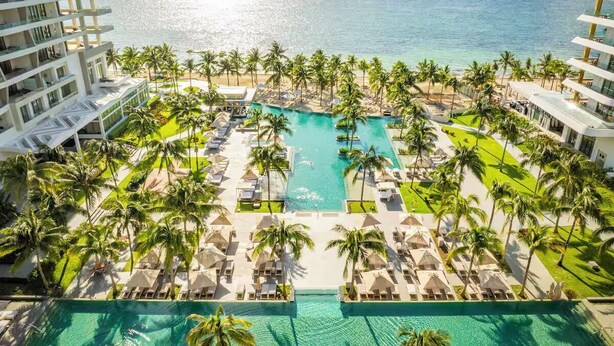 Ein Resort mit einem zentral gelegenen Pool, umgeben von Palmen und Liegestühlen, mit Blick auf einen Strand und den Ozean.