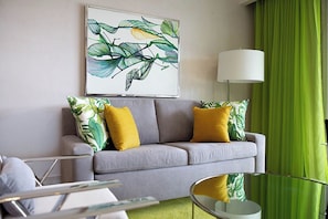 Una sala de estar moderna con un sofá gris, cojines amarillos, un mural de plantas verdes, una lámpara de pie blanca y una cortina verde.