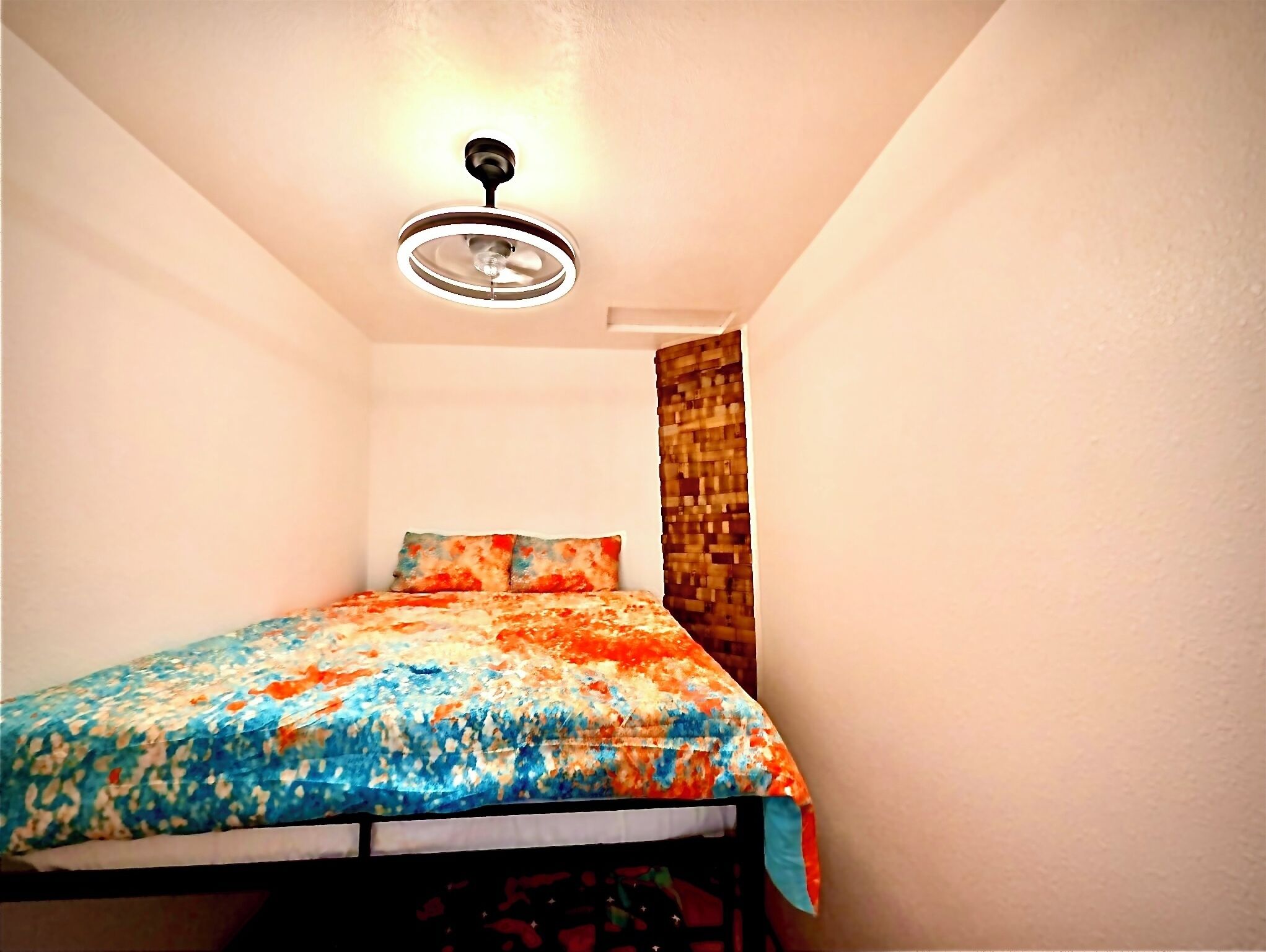 A small, well-lit bedroom with a bed, a ceiling fan, and a window with a curtain.