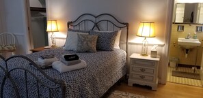 A neatly made bed with patterned bedding, a bedside table with a lamp, a small drawer unit, and a bathroom with a sink and mirror. - 3-bedroom house in lovely Halifax for quiet, restful stays (Halifax)