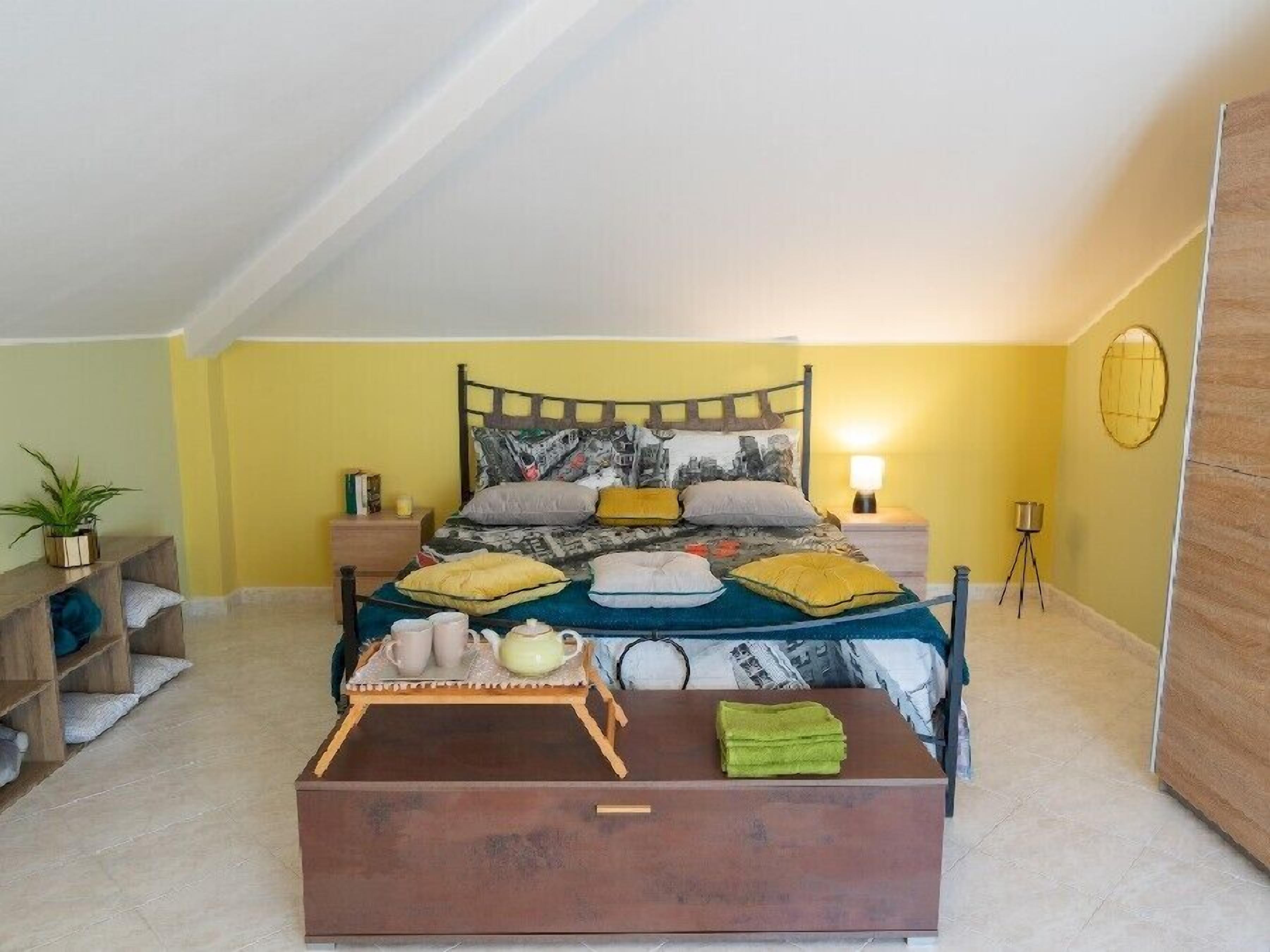 Un dormitorio con una cama, mesitas de noche, una lámpara, una lámpara de pie, una planta en maceta, un banco de madera y una pequeña mesa con tetera y tazas.