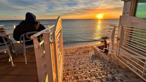 Un escalier menant à une plage offrant une vue sur le coucher de soleil, une personne assise sur une chaise, et une pancarte indiquant « Le Buno ».