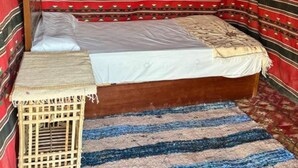 A single bed with a wooden frame, a white mattress, and a red and black striped blanket. There is a woven basket beside the bed and a blue and beige striped rug on the floor.