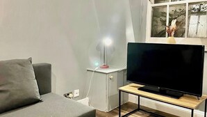 A modern living room with a grey sofa, a wooden coffee table with a remote control and a potted plant, a TV on a wooden stand, and a small side table with a lamp. - Cosy Flat Near Kings Cross (London)