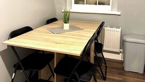 A small, neatly arranged dining area with a wooden table, four black chairs, a potted plant, and a window with frosted glass panes. - Cosy Flat Near Kings Cross (London)