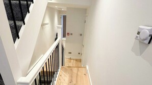 A staircase with black railings leading to a landing with a white wall and a thermostat on the wall. - Cosy Flat Near Kings Cross (London)