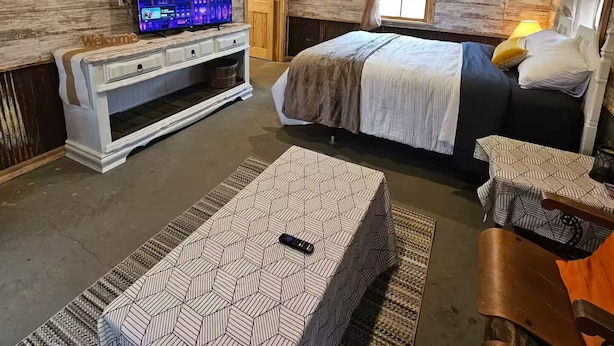 A rustic room with a wooden ceiling, a bed with a patterned bedspread, a TV on a wooden cabinet, and a window with curtains. - Relaxing Fun Bunk house 2 (Weyerhaeuser)