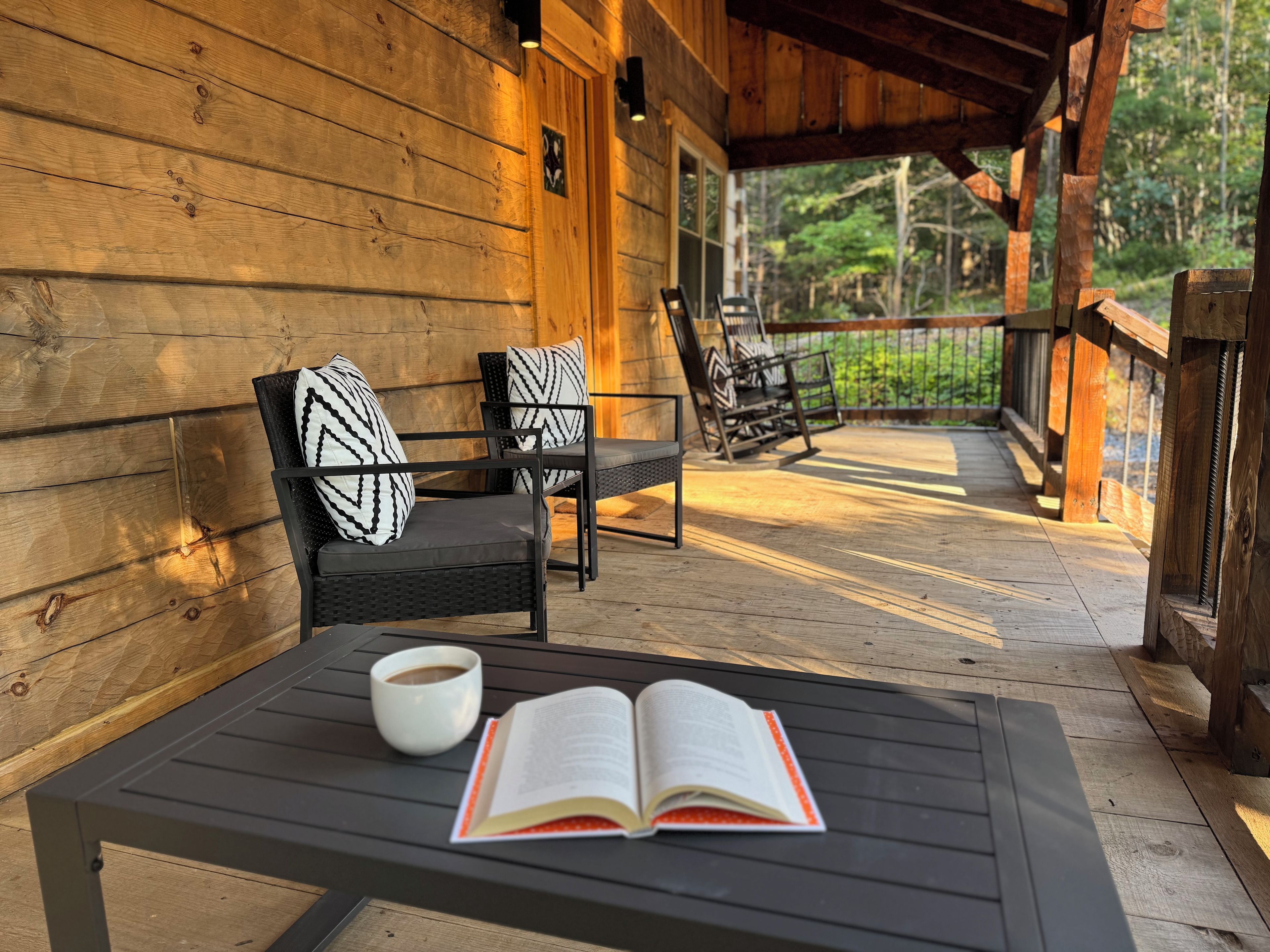 A wooden deck with outdoor furniture, including a table with a book and a cup, and a bench with patterned cushions.