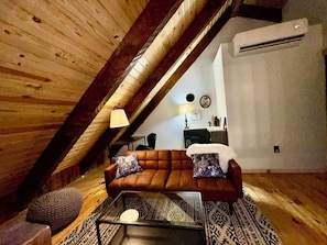 Habitación estilo loft con techo de madera, sofá con cojines estampados, una mesa de centro de vidrio y alfombra con diseño.