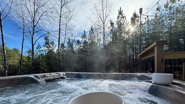 Un jacuzzi con muelle de madera y una taza de café, rodeado de árboles y un cielo despejado.
