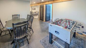 A room with a foosball table, a dining table with chairs, and a sofa in the background.