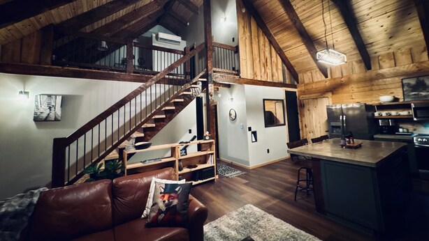 A spacious loft with a high ceiling, wooden beams, a staircase, a kitchen area, and a living room with a leather couch.