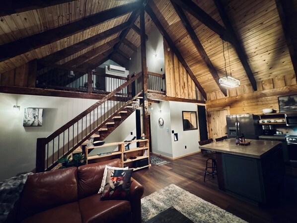 A spacious loft with a high ceiling, wooden beams, a staircase, a kitchen area, and a living room with a leather couch.