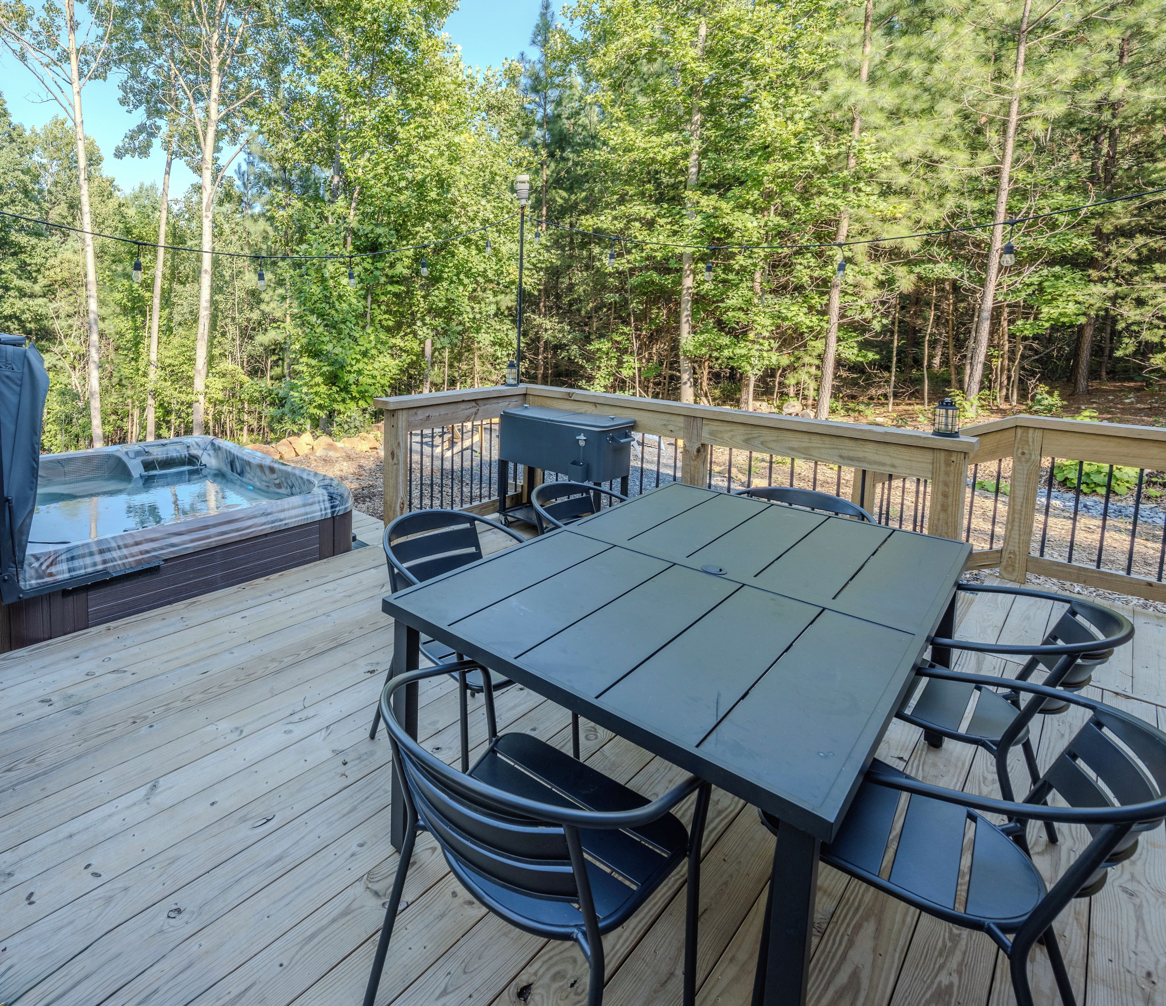 A wooden deck with a table and chairs, a hot tub, and a railing overlooking a forested area.