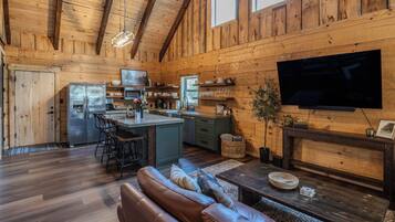 A cozy cabin interior with a wooden interior, a kitchen with a refrigerator, a dining area with a sofa, and a flat-screen TV mounted on the wall.