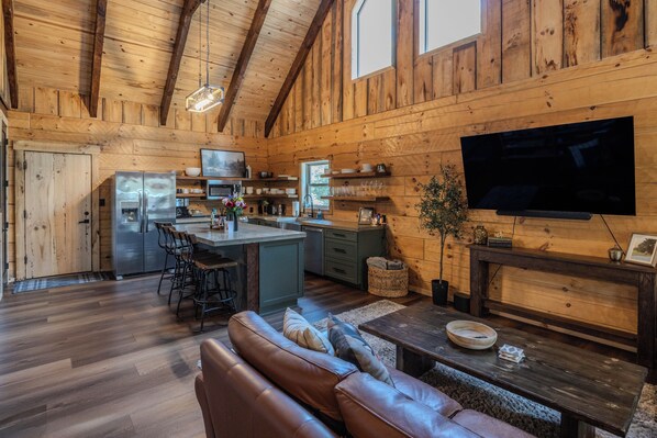 Interior acogedor de una cabaña con detalles de madera, una cocina con refrigerador, un comedor con sofá y un televisor de pantalla plana montado en la pared.