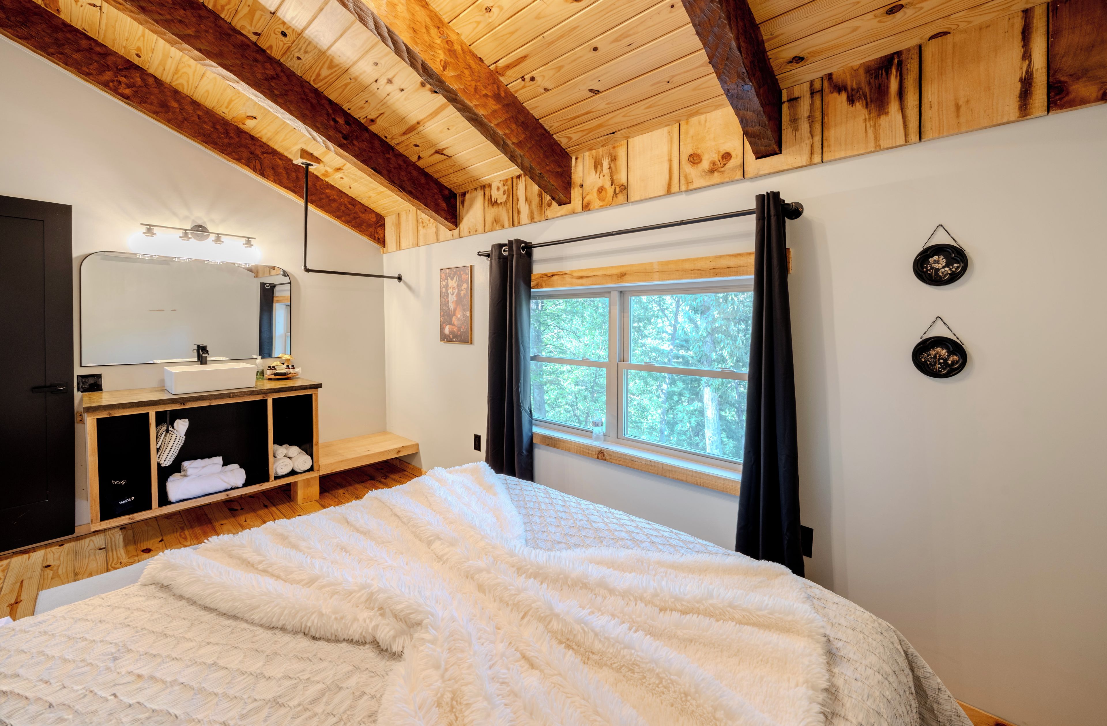 Un dormitorio con techo de madera, una cama con manta blanca y esponjosa, un aparador de madera, un espejo y una ventana con cortinas.