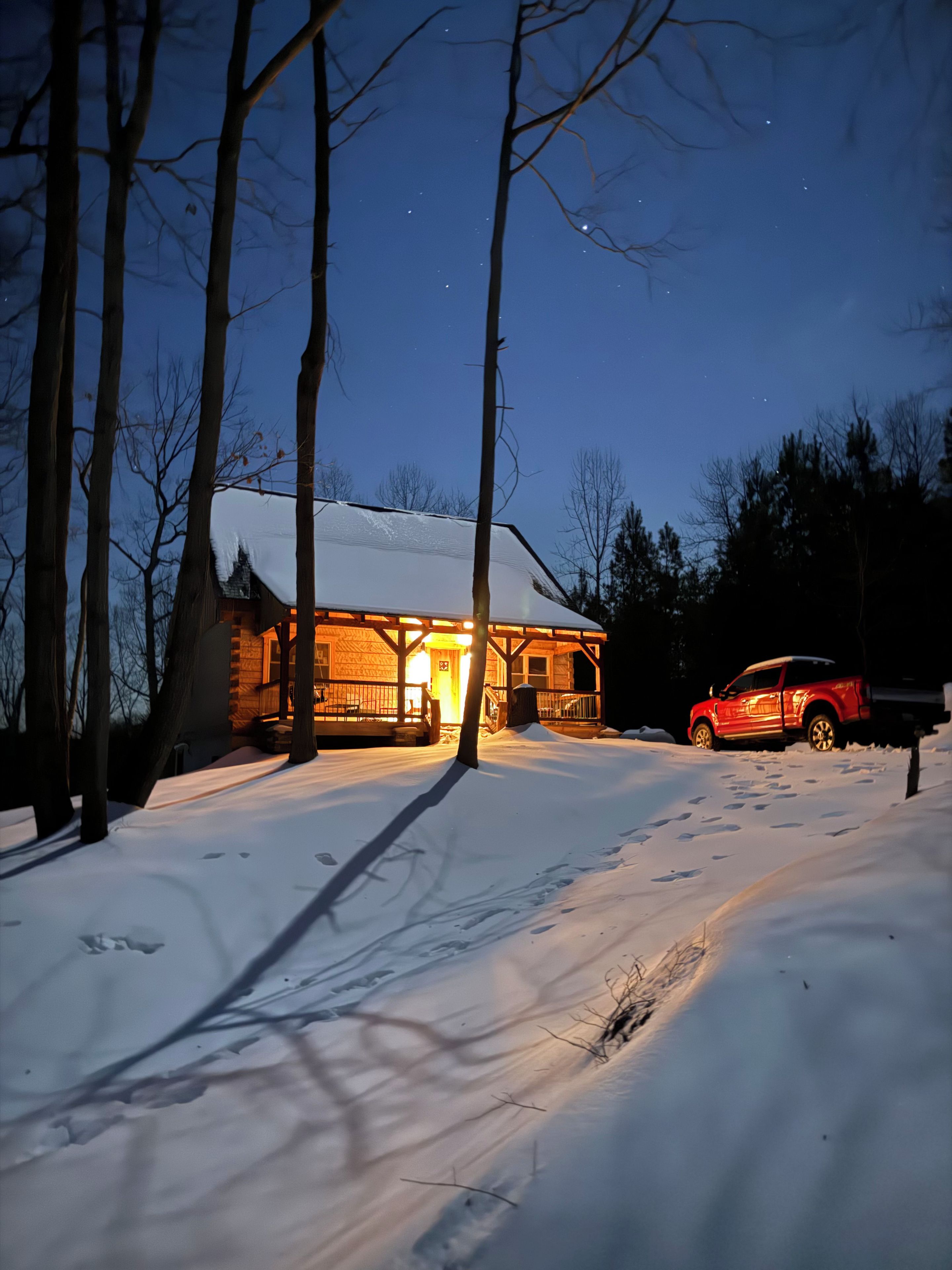 A cabin with a snow-covered roof, a wooden deck, and a red vehicle parked in front.