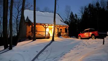A cabin with a snow-covered roof, a wooden deck, and a red vehicle parked in front.
