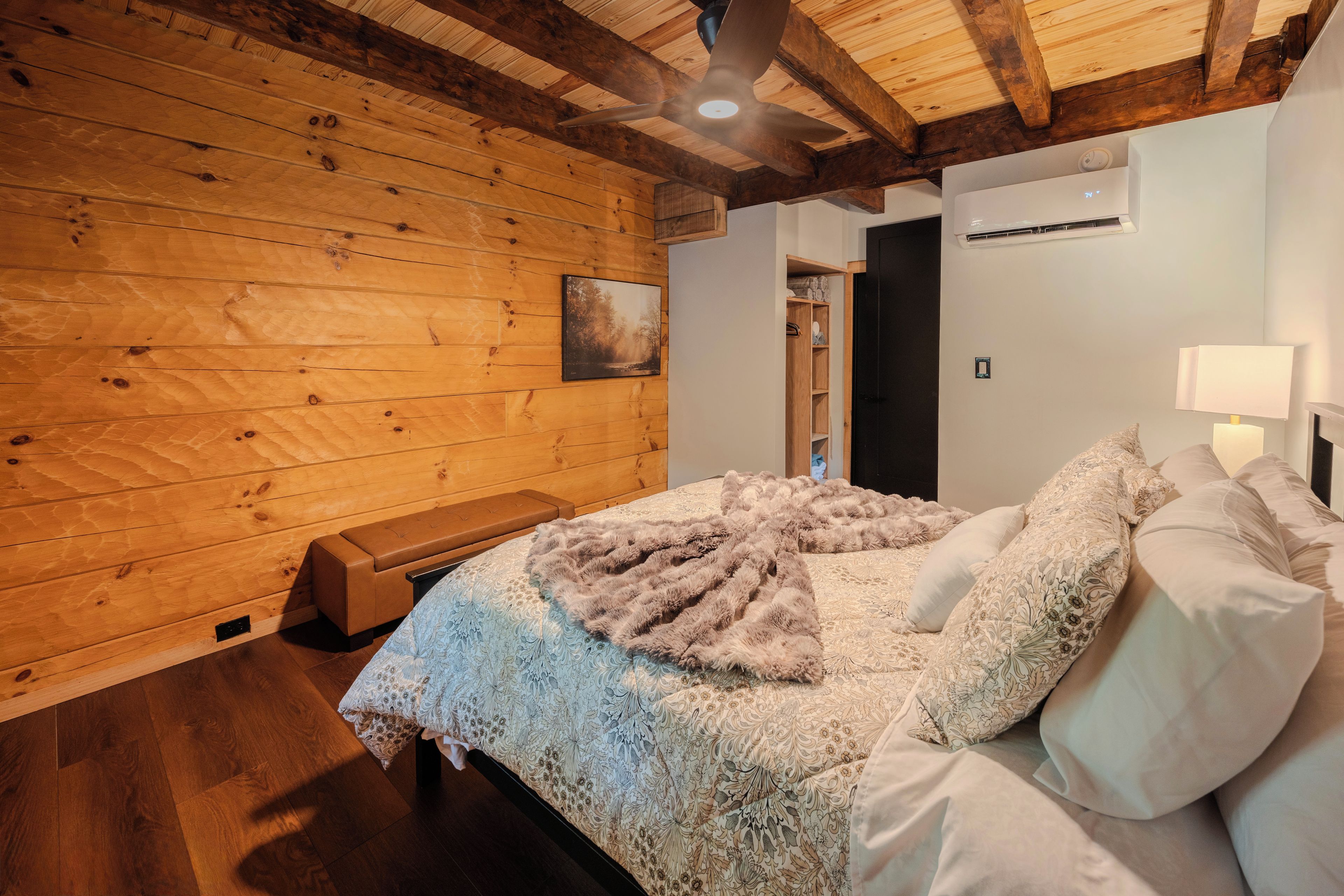 A bedroom with a wooden ceiling, a bed with patterned bedding, a bench, and an air conditioning unit.