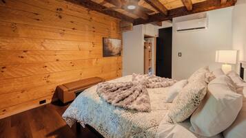 A bedroom with a wooden ceiling, a bed with patterned bedding, a bench, and an air conditioning unit.
