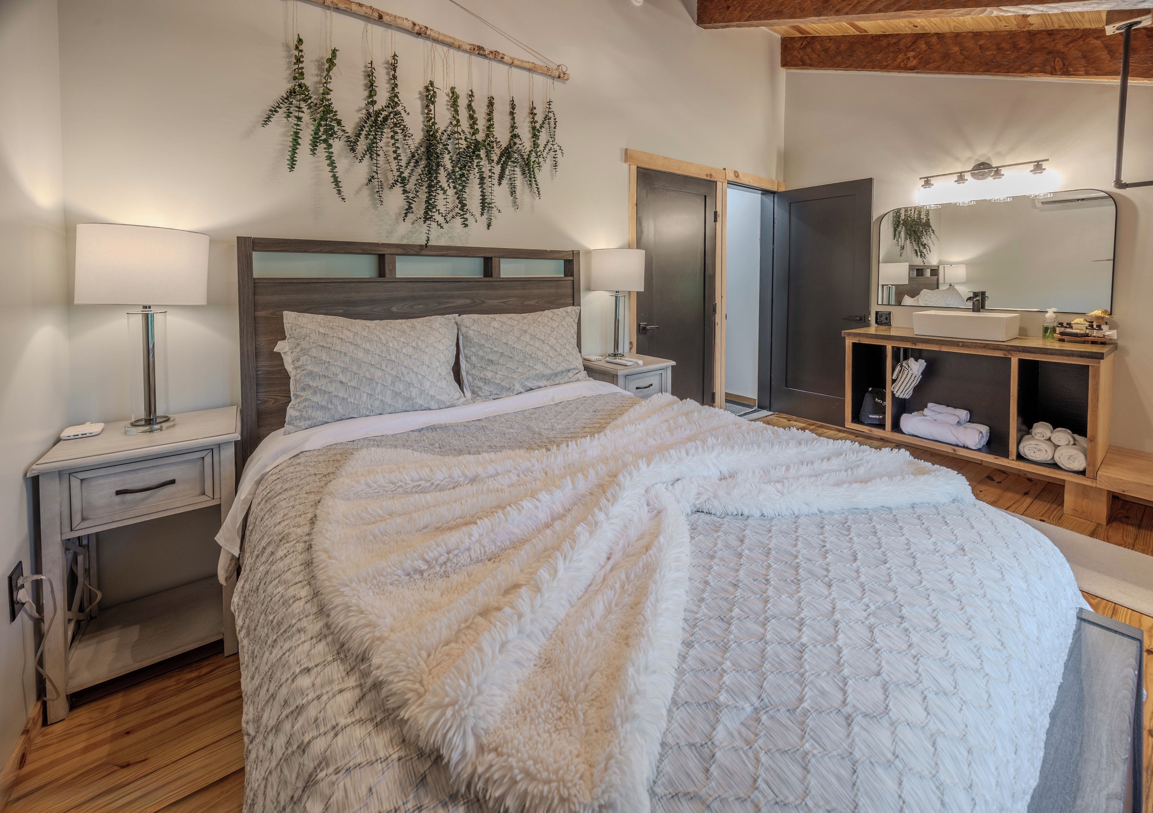 A bedroom with a bed, bedside tables, a nightstand, a dresser, a mirror, and a hanging plant.