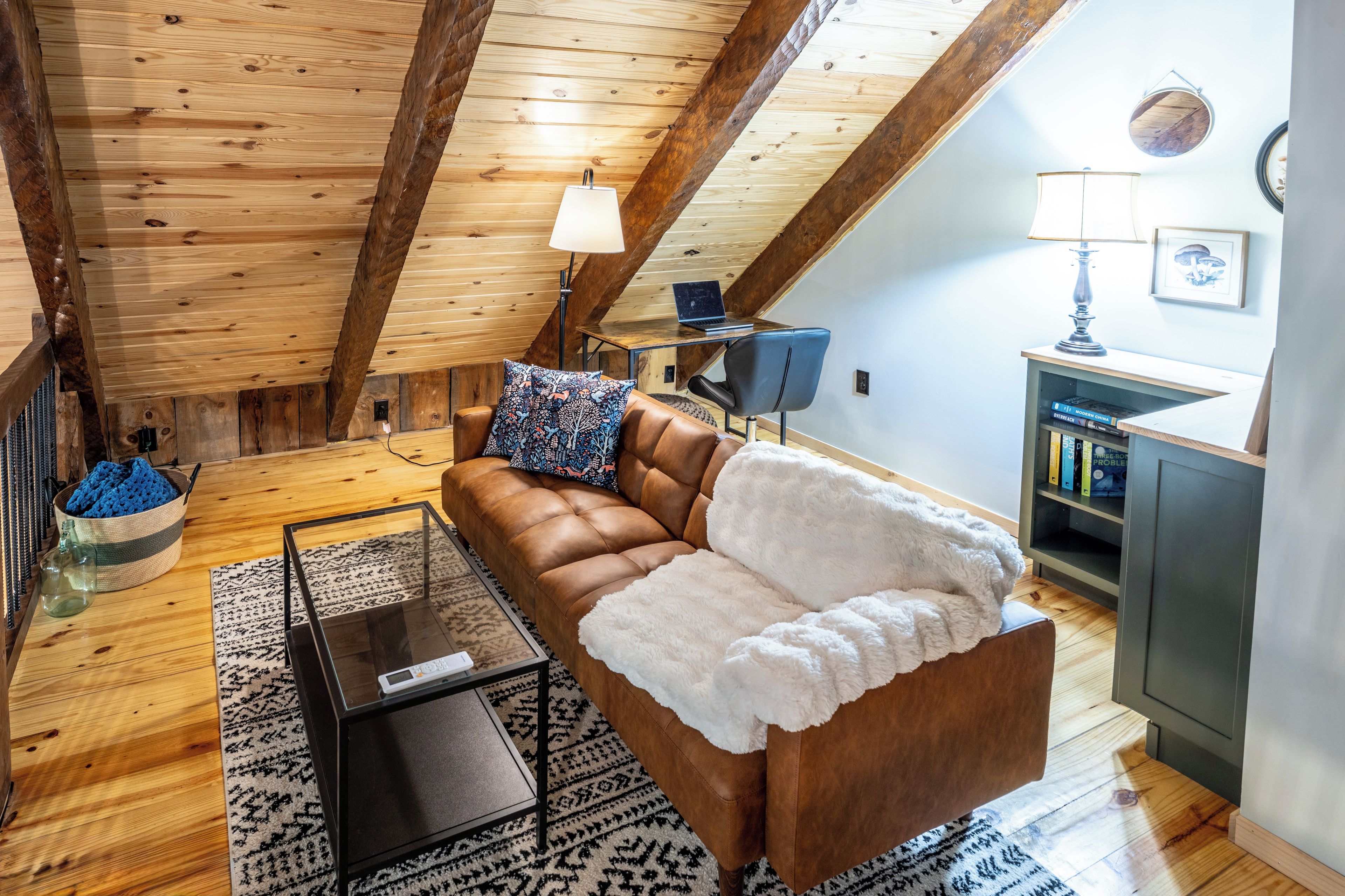 A cozy loft bedroom with a wooden ceiling, a brown leather sofa, a glass coffee table, and a patterned rug.