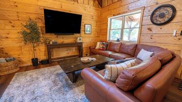 A cozy cabin interior with a leather sofa, a flat-screen TV mounted on the wall, and a rustic wooden floor.
