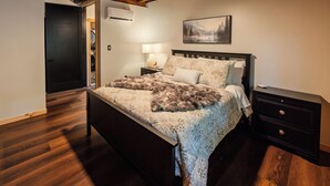 Un dormitorio con techo de madera, una cama con ropa de cama estampada, una mesita de noche con lámpara y un cuadro en la pared.